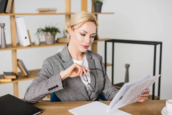 Geschäftsfrau mit Brille in der Hand liest Zeitung am Arbeitsplatz — Stockfoto