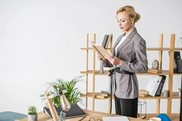 Впевнена бізнес-леді в костюмі з блокнотом, що стоїть в офісі — стокове фото