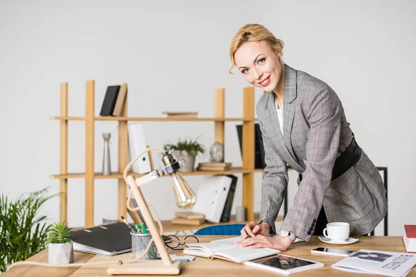 Porträt einer lächelnden Geschäftsfrau, die in die Kamera blickt, während sie am Arbeitsplatz Notizen in Notizbuch macht — Stockfoto