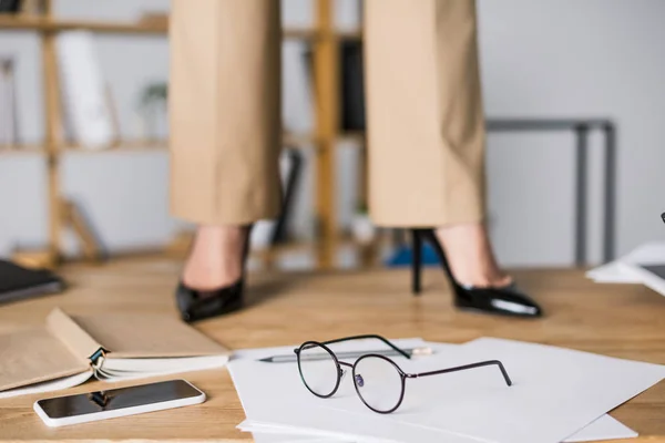 Cropped shot of businesswoman standing near smartphone, papers and eyeglasses on floor — Stock Photo
