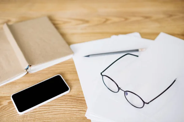 Foyer sélectif du smartphone avec écran vierge, lunettes et papiers sur table en bois — Photo de stock