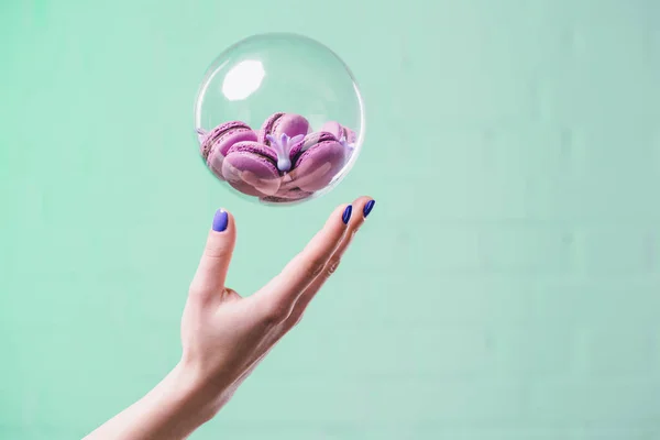 Cropped shot of woman throwing up glass ball with macarons on turquoise — Stock Photo