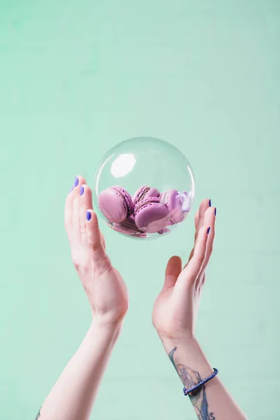 Cropped shot of woman throwing up glass ball with delicious macarons on turquoise — Stock Photo