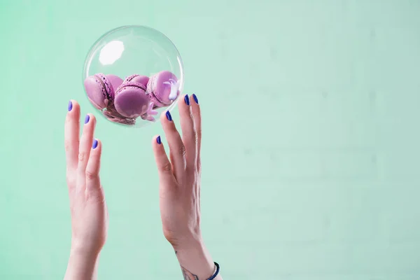 Cropped shot of woman throwing up glass ball with tasty macarons on turquoise — Stock Photo