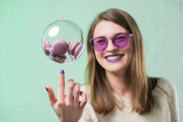 Happy young woman in pink eyeglasses touching glass ball of macarons — Stock Photo