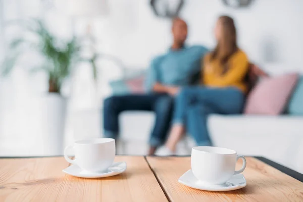 Multicultural couple holding hands on sofa with cups of coffee on foreground — Stock Photo