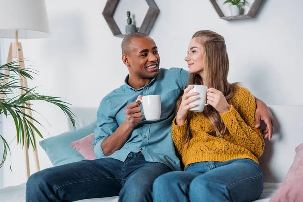 Мультикультурная пара сидит на диване с кофе и смотрит друг на друга — стоковое фото