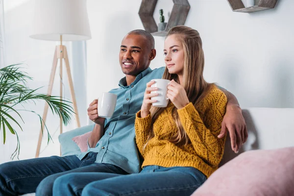 Casal multicultural sentado no sofá com café e olhando para longe — Fotografia de Stock