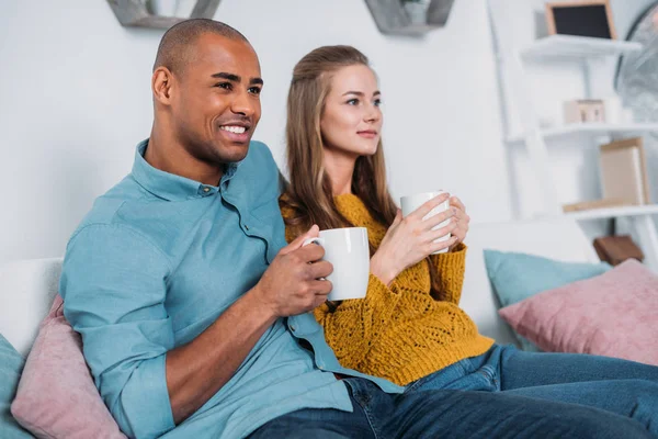 Multikulturelles Paar sitzt mit Kaffee auf Sofa und schaut weg — Stockfoto