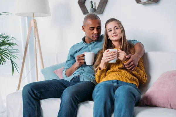 Multicultural couple hugging and sitting on sofa with coffee — Stock Photo