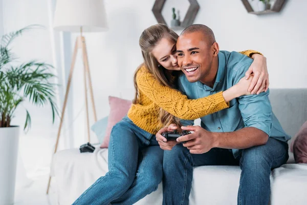 Namorada abraçando o namorado afro-americano jogando videogame em casa — Fotografia de Stock