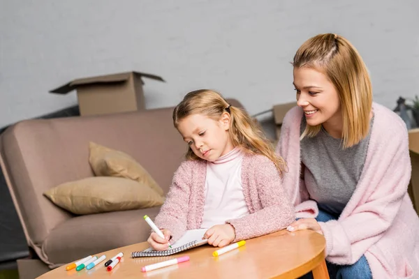 Glückliche Mutter betrachtet Tochter Zeichnung mit Filzstiften am Tisch — Stockfoto