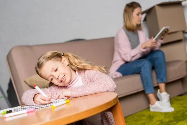 Cute little child drawing at table while mother using digital tablet on sofa — Stock Photo