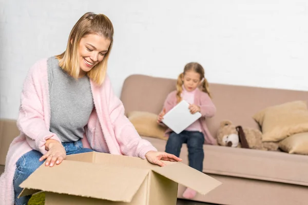 Mujer sonriente embalaje caja de cartón mientras que la pequeña hija usando tableta digital - foto de stock