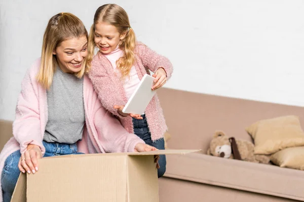 Feliz madre e hija con caja de cartón de embalaje de tableta digital durante la reubicación - foto de stock