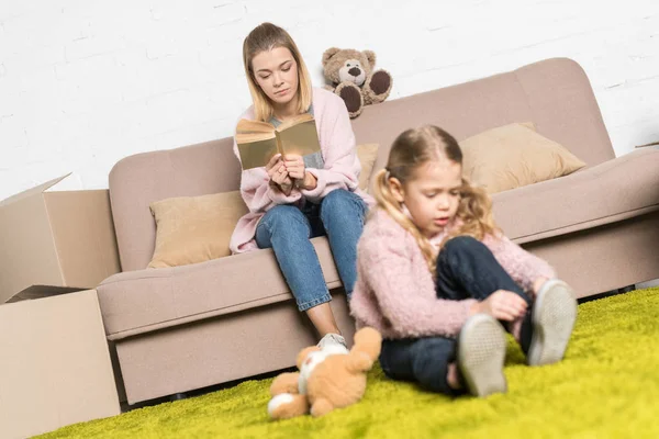 Kind spielt mit Teddybär auf Teppich, während Mutter Buch auf Sofa liest — Stockfoto