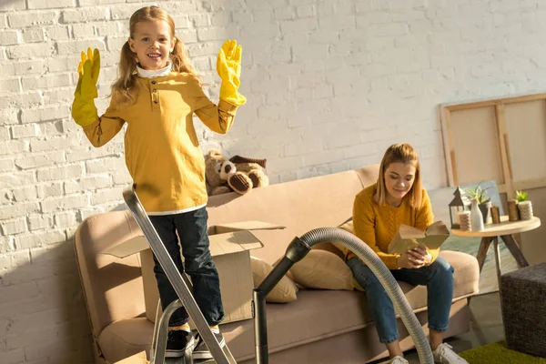 Child in rubber gloves smiling at camera while mother reading book on sofa during relocation — Stock Photo