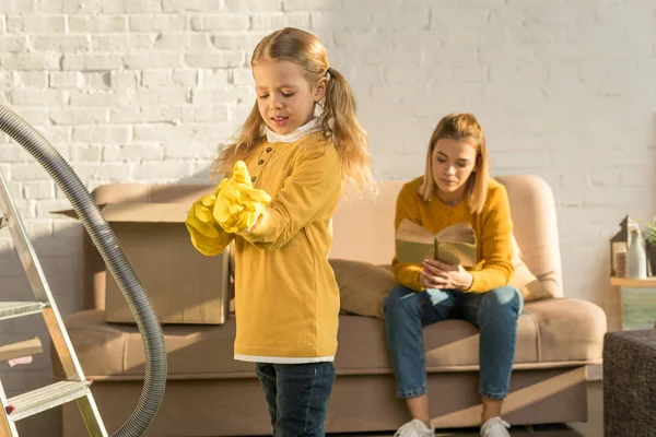 Kind trägt Gummihandschuhe beim Putzen und Mutter liest Buch auf dem Sofa nach dem Umzug — Stockfoto