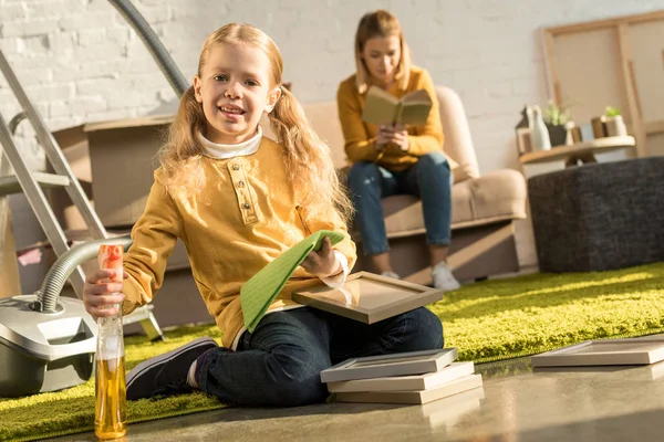 Cute little child cleaning frames and smiling at camera while mother reading book after relocation — Stock Photo