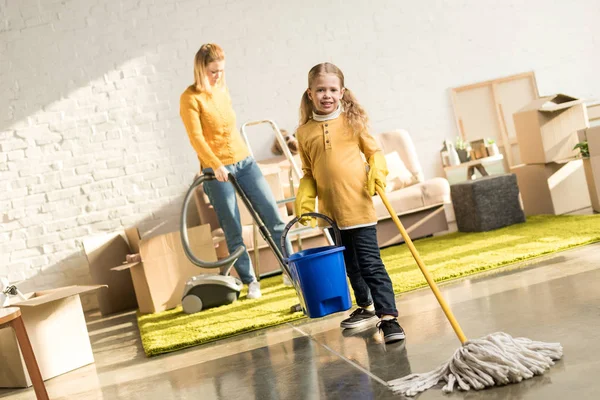 Madre e hija con fregona y aspiradora sala de limpieza mientras se muda a casa - foto de stock