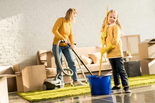 Madre e hija con fregona y aspiradora sala de limpieza después de la reubicación - foto de stock