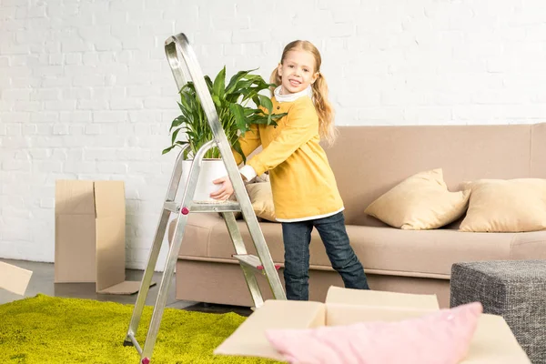 Adorable niño pequeño sosteniendo planta de interior y sonriendo a la cámara mientras se muda a casa - foto de stock