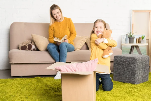 Heureuse petite fille étreignant ours en peluche et regardant la caméra tout en souriant mère lecture livre sur canapé — Photo de stock