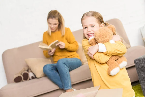 Mignonne petite fille étreignant ours en peluche et souriant à la caméra tandis que la mère lit le livre sur le canapé — Photo de stock