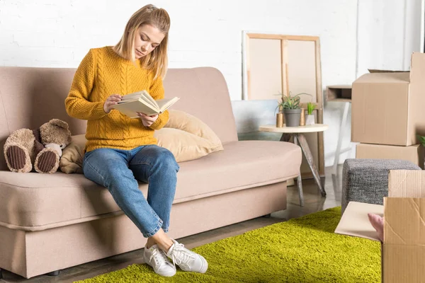Attractive young woman sitting on sofa and reading book during relocation — Stock Photo