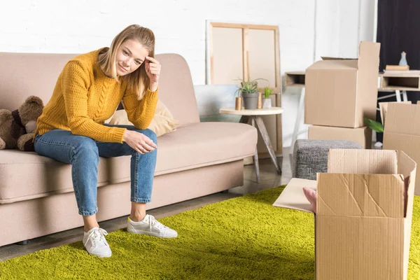 Smiling young woman sitting on sofa while packing cardboard boxes during relocation — Stock Photo