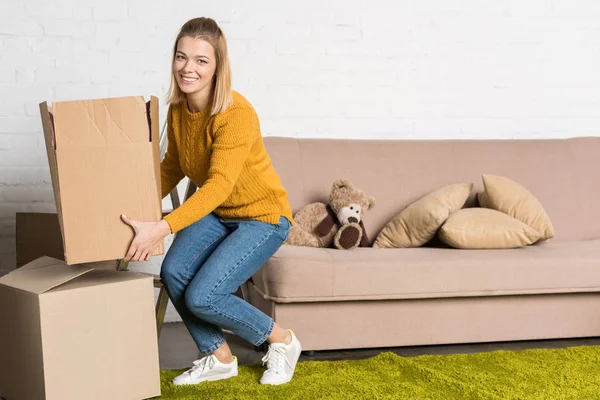Woman holding cardboard box and smiling at camera while relocating — Stock Photo