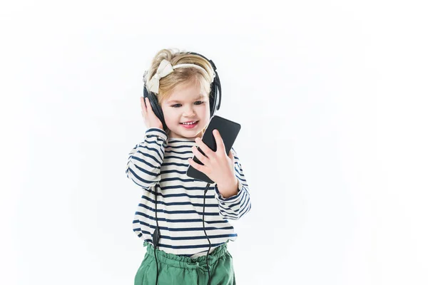 Прелестный маленький ребенок слушает музыку с наушниками и смартфоном изолированы на белом — стоковое фото