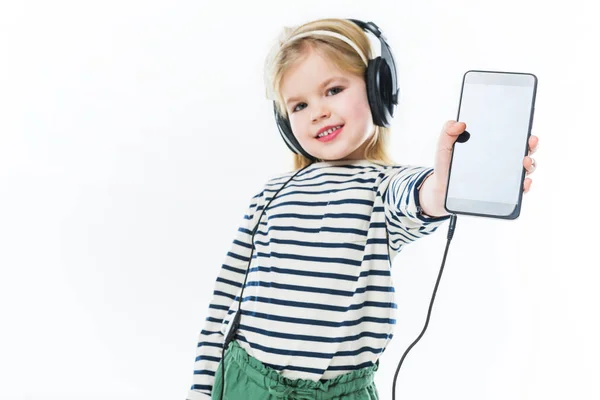 Criança pequena feliz ouvir música com fones de ouvido e mostrando smartphone isolado no branco — Fotografia de Stock