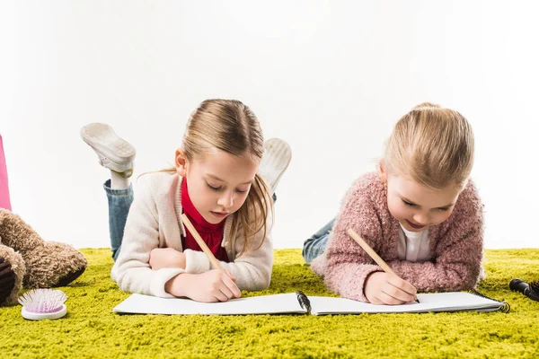 Hermanas pequeñas enfocadas dibujando con lápices de color juntos en el suelo aislado en blanco - foto de stock