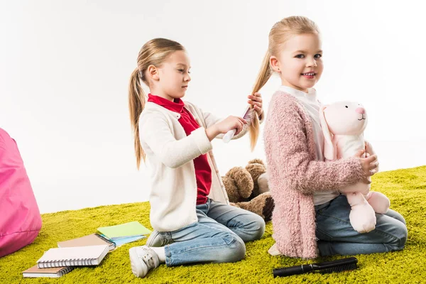 Beautiful child brushing hair of sister while she sitting on floor with toy bunny isolated on white — Stock Photo