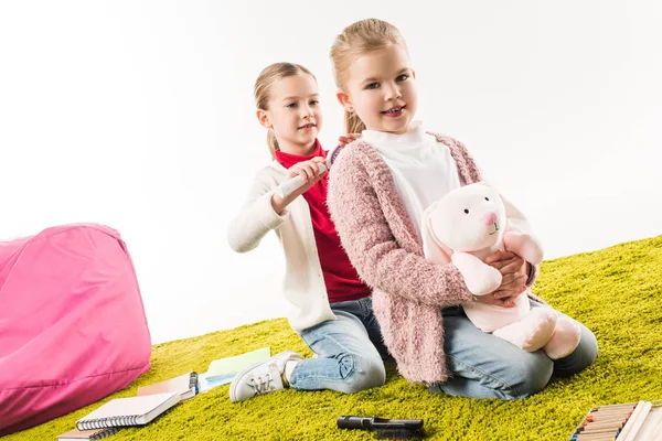 Eine Schwester bürstet der anderen die Haare, während sie auf dem Boden sitzt, mit Spielzeughase isoliert auf weiß — Stockfoto