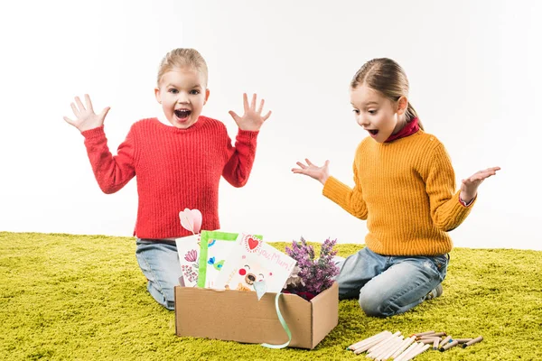 Aufgeregte kleine Schwestern mit einer Kiste voller Materialien für die Kunst sitzen auf dem Boden isoliert auf weiß — Stockfoto