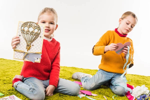 Hermanitas enfocadas haciendo tarjetas de felicitación mientras están sentadas en el piso aisladas en blanco - foto de stock