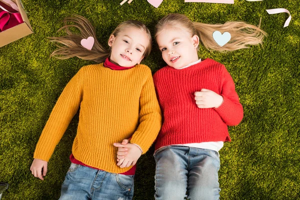 Vista superior de hermanas pequeñas acostadas en la alfombra y cogidas de la mano rodeadas de corazones - foto de stock