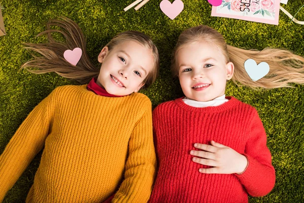 Top view of happy little sisters lying on carpet surrounded with hearts — Stock Photo