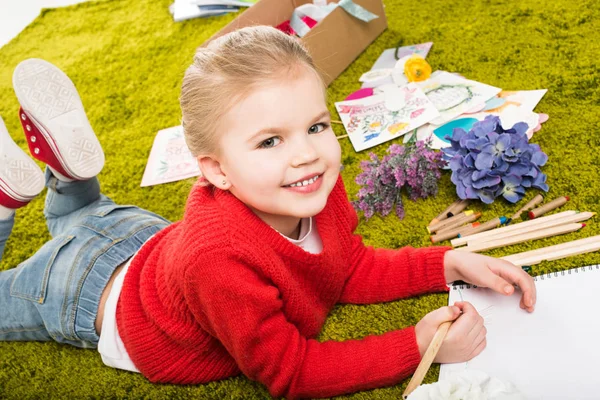 Усміхнений малюнок маленької дитини з кольоровими олівцями на зеленому м'якому килимі — стокове фото