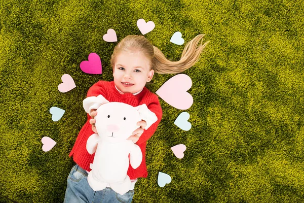 Vista superior de un niño feliz rodeado de corazones acostados en una alfombra verde y suave con conejo de juguete - foto de stock