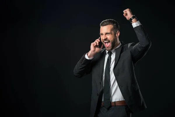 Hombre de negocios emocionado en la chaqueta celebrando mientras habla en el teléfono inteligente, aislado en negro - foto de stock