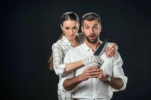 Шокований чоловік тримає доларові банкноти, а жінка тримає ніж на шиї, ізольований на чорному — стокове фото