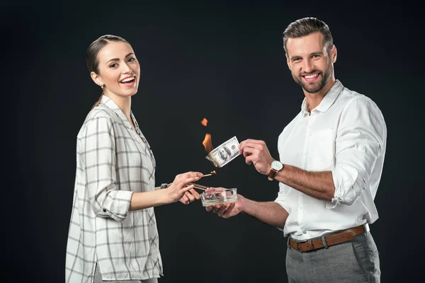 Sonriente hombre y mujer quemando billetes de dólar, aislados en negro - foto de stock