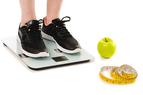 Cropped shot of woman standing on scales with apple and measuring tape lying on floor isolated on white — Stock Photo