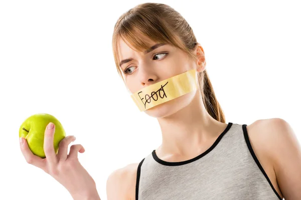 Giovane donna con nastro adesivo con striked attraverso parola cibo che copre bocca guardando mela in mano isolato su bianco — Foto stock