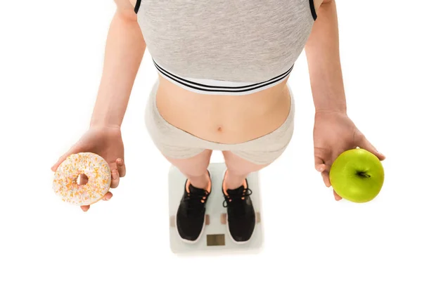 Cropped shot of slim woman with apple and doughnut standing on scales isolated on white — Stock Photo