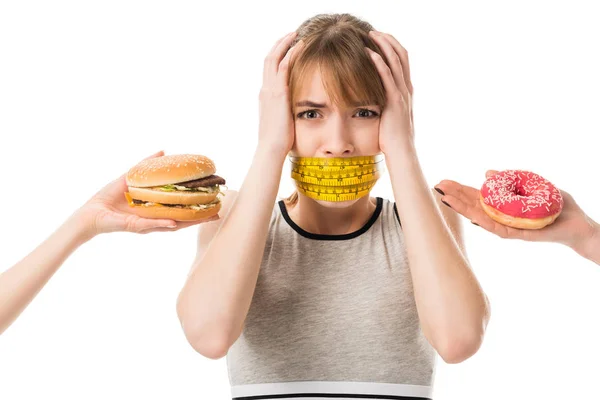 Jovem mulher com a boca amarrada em fita métrica gritando enquanto as pessoas dando-lhe junk food isolado no branco — Fotografia de Stock