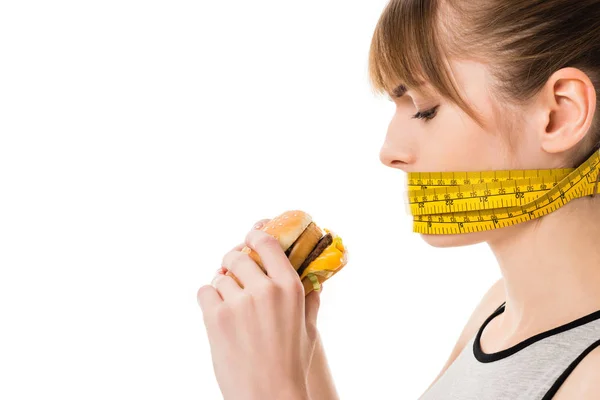 Mulher com a boca amarrada em fita métrica tentando comer hambúrguer isolado em branco — Fotografia de Stock